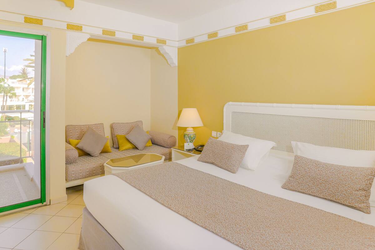 A hotel room with a bed, a sofa, a table, and a lamp. There is a window with a view of palm trees and buildings.