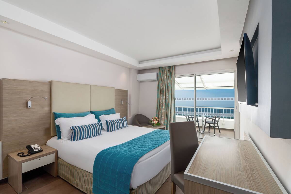 A hotel room with a bed, bedside table, desk, and a view of the sea.