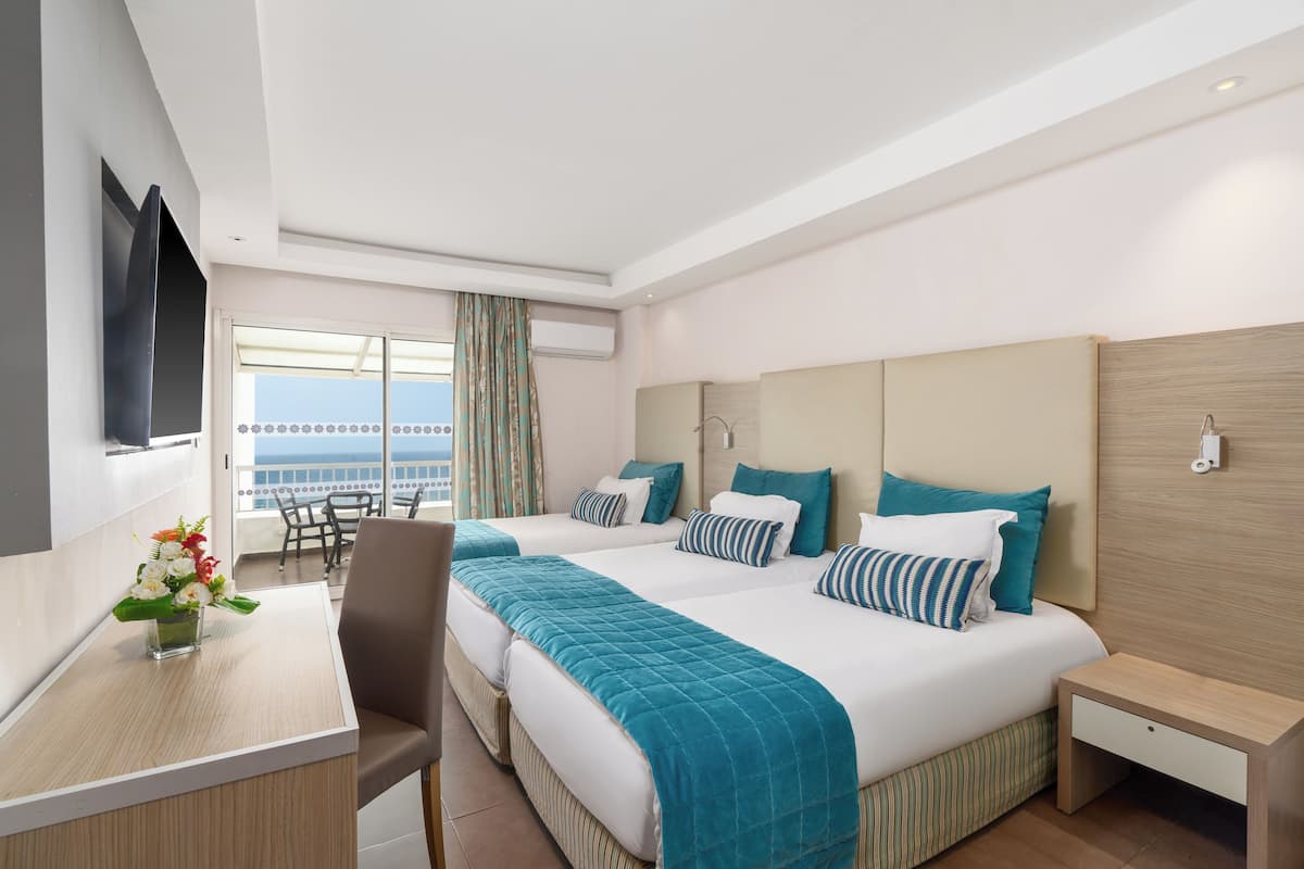 A hotel room with two beds, a desk, a chair, a TV, and a balcony with a view.