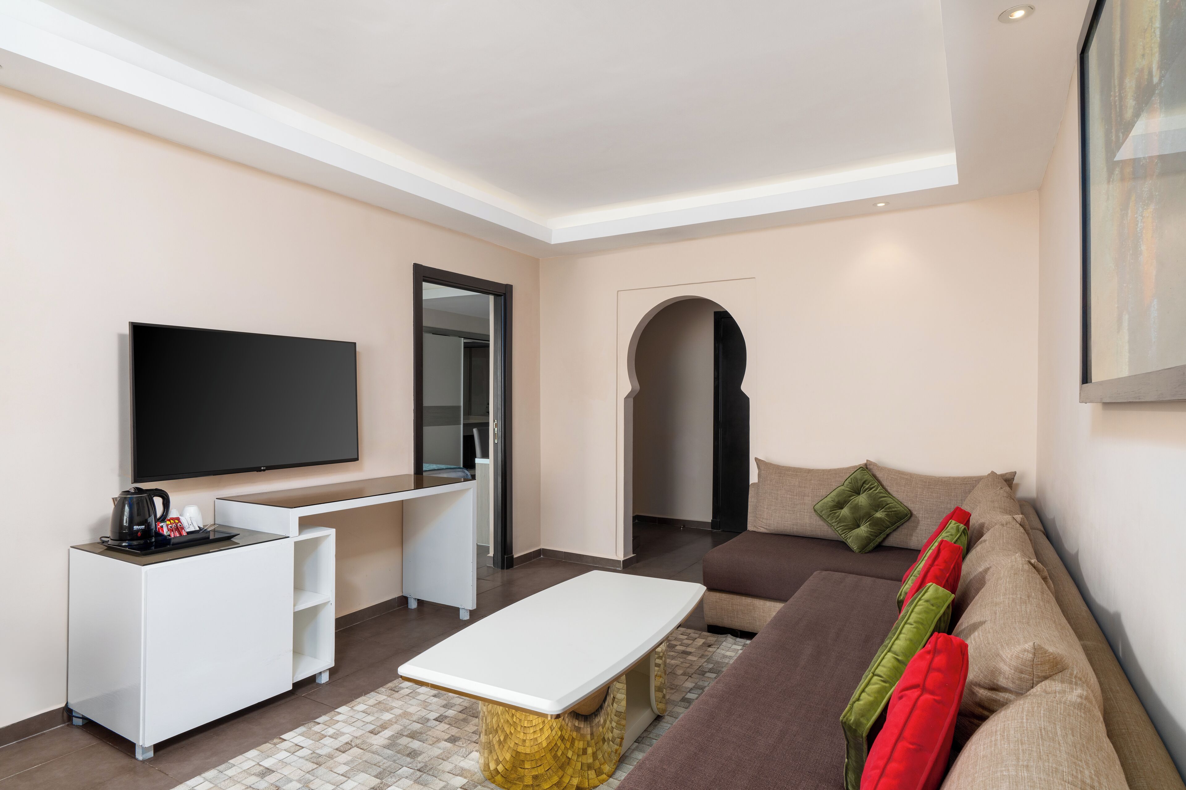 Una sala moderna con una televisión de pantalla plana, una mesa de centro blanca, un sofá marrón y un pequeño escritorio blanco con una cafetera.