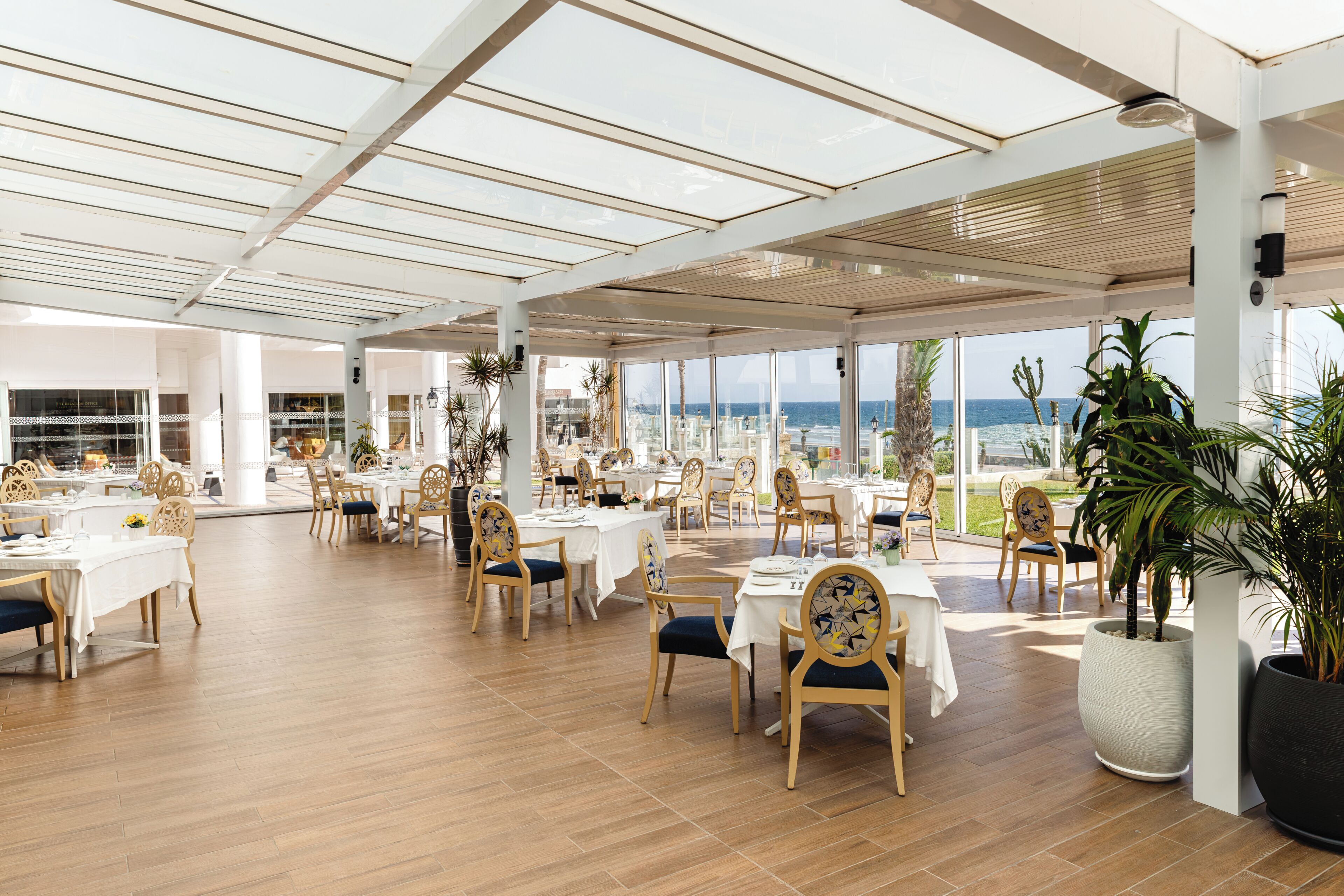 A spacious indoor dining area with multiple tables and chairs, large windows offering a view of the sea, and a high ceiling with natural light.