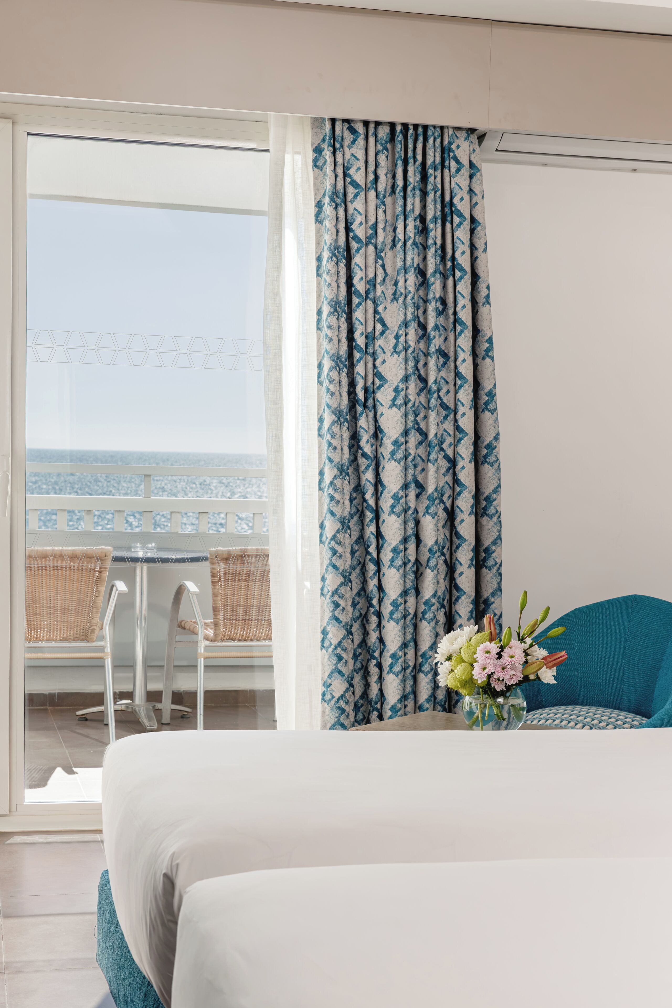Hotelkamer met een bed, uitzicht op de zee, een balkon met rieten stoelen en een tafel met een bloevas.
