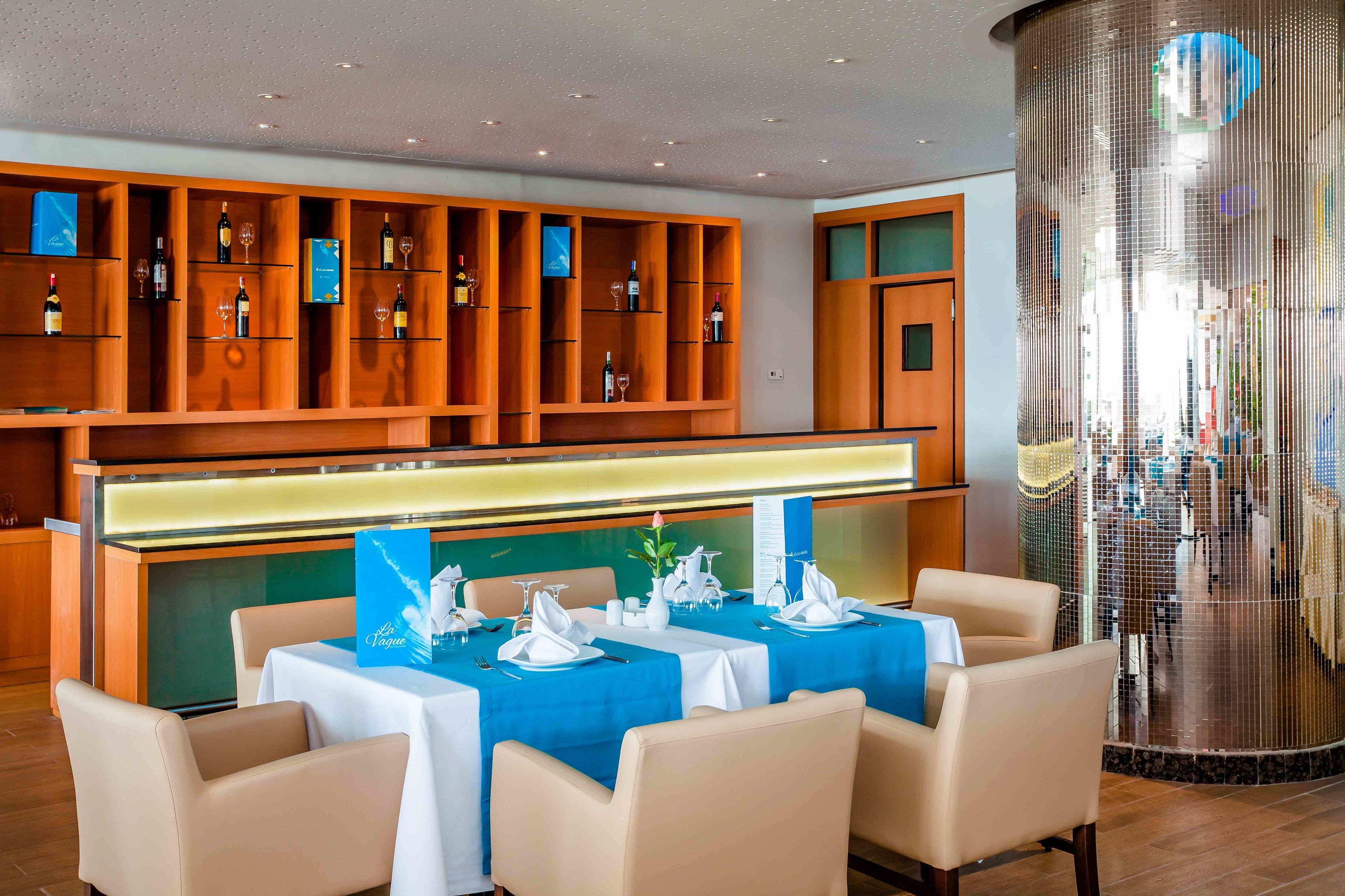 Een restaurant met houten schappen waarop wijnflessen en glazen staan, tafels gedekt met witte tafelkleden en servetten, en een grote metalen kolom op de achtergrond.