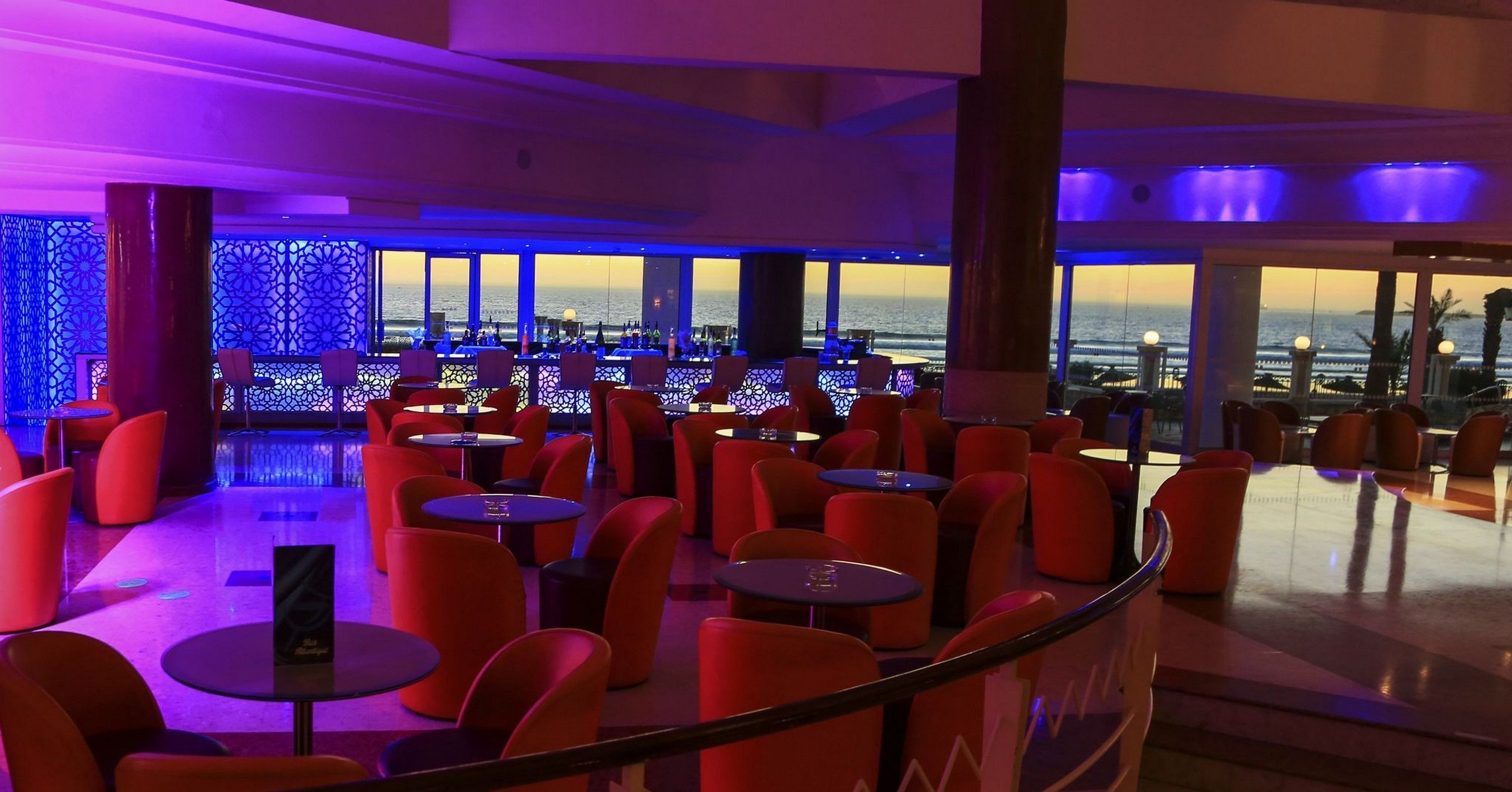 A modern lounge area with red chairs and tables, large windows offering a view of the sea, and a decorative blue-lit wall.