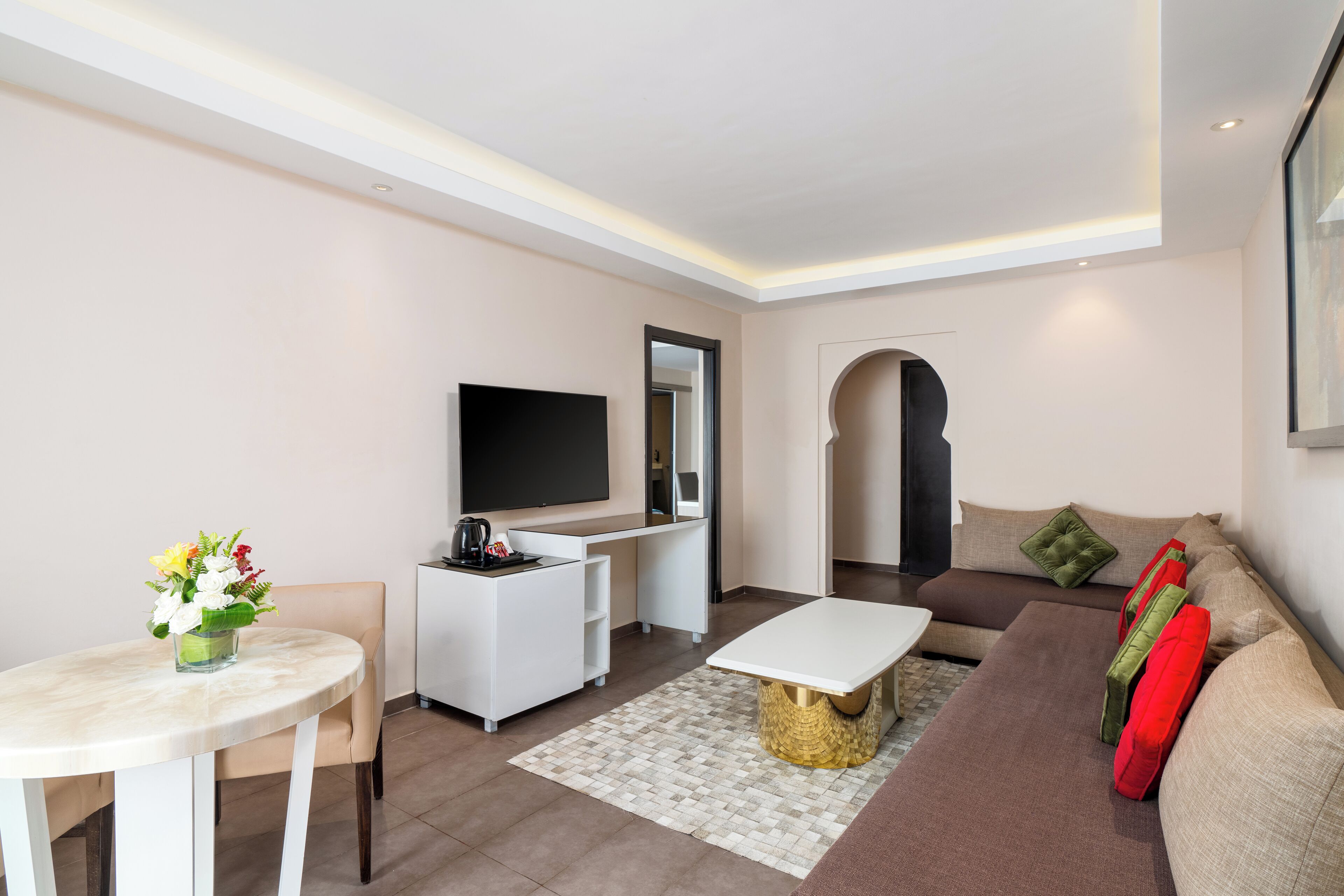 A modern living room with a flat-screen TV, a white desk, a round table with a vase of flowers, a sofa with cushions, and a patterned rug.