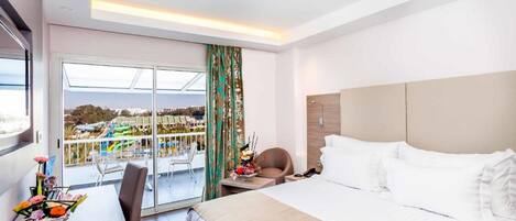 A hotel room with a large bed, a desk with a chair, a small table with a vase of flowers, and a balcony with a view of the city.
