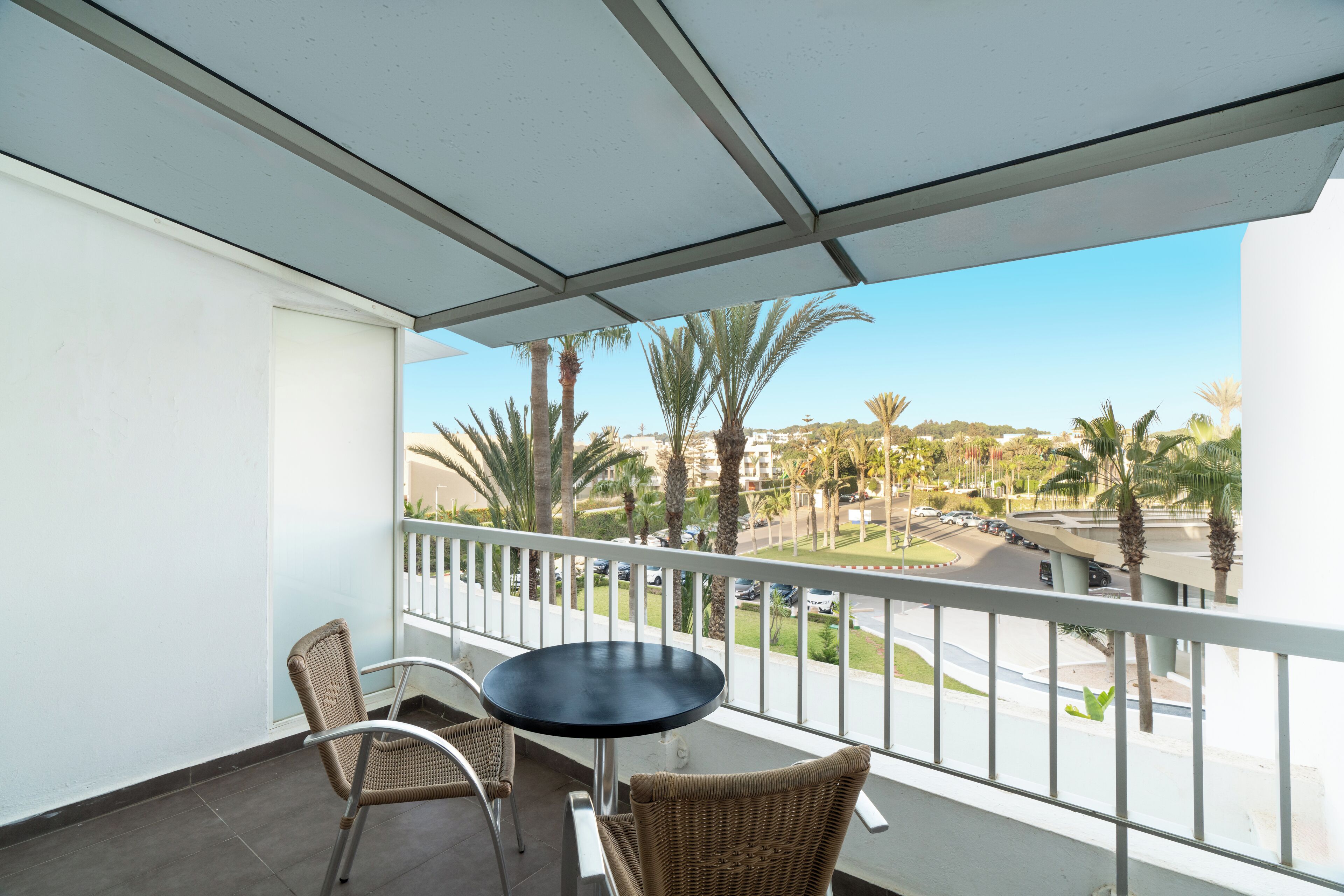 A balcony with a table and chairs, overlooking a street with palm trees and a clear sky.