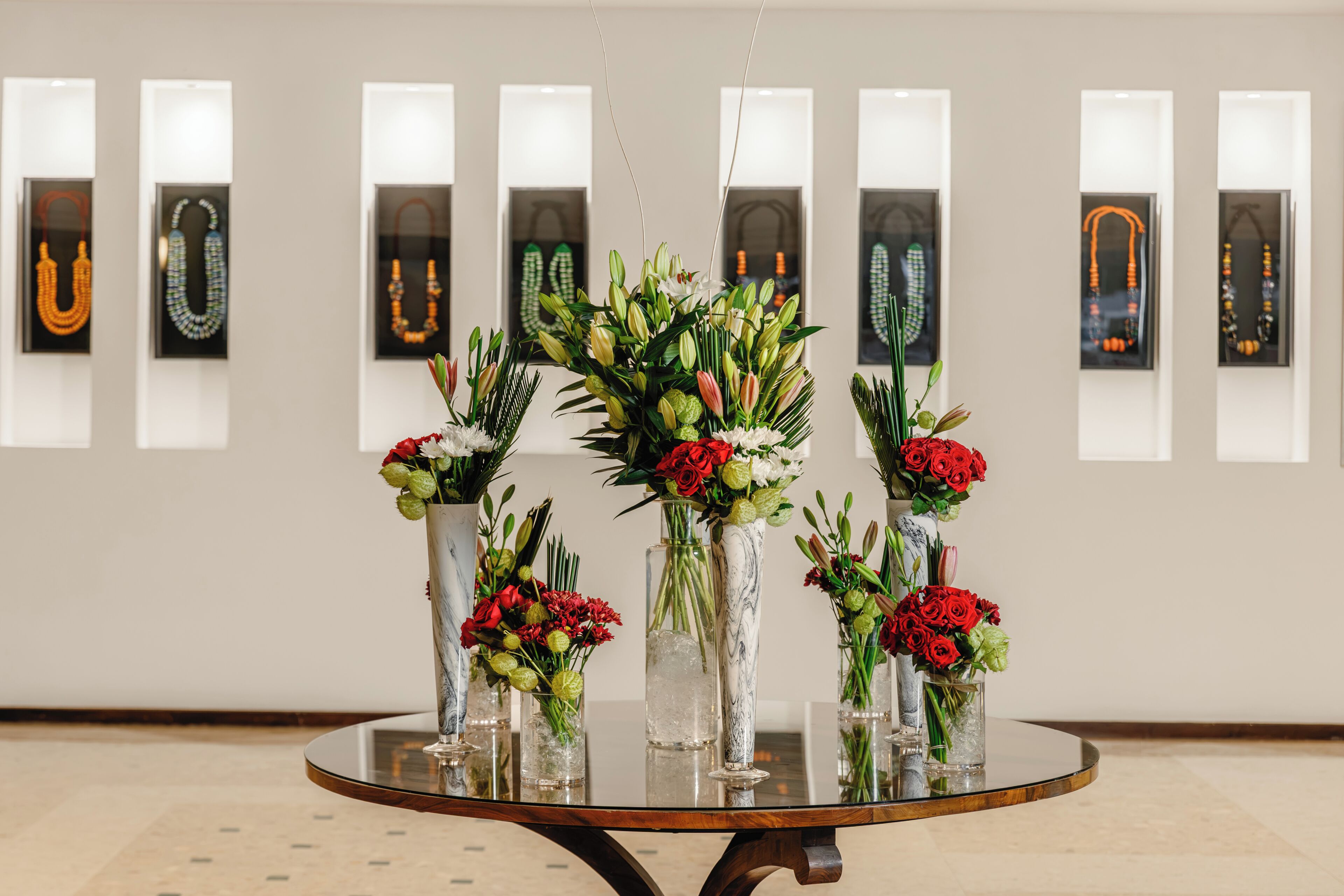 Ein runder Glastisch mit floralen Dekorationen in weißen Vasen, vor einer Wand mit gerahmten Schmuckpräsentationen.