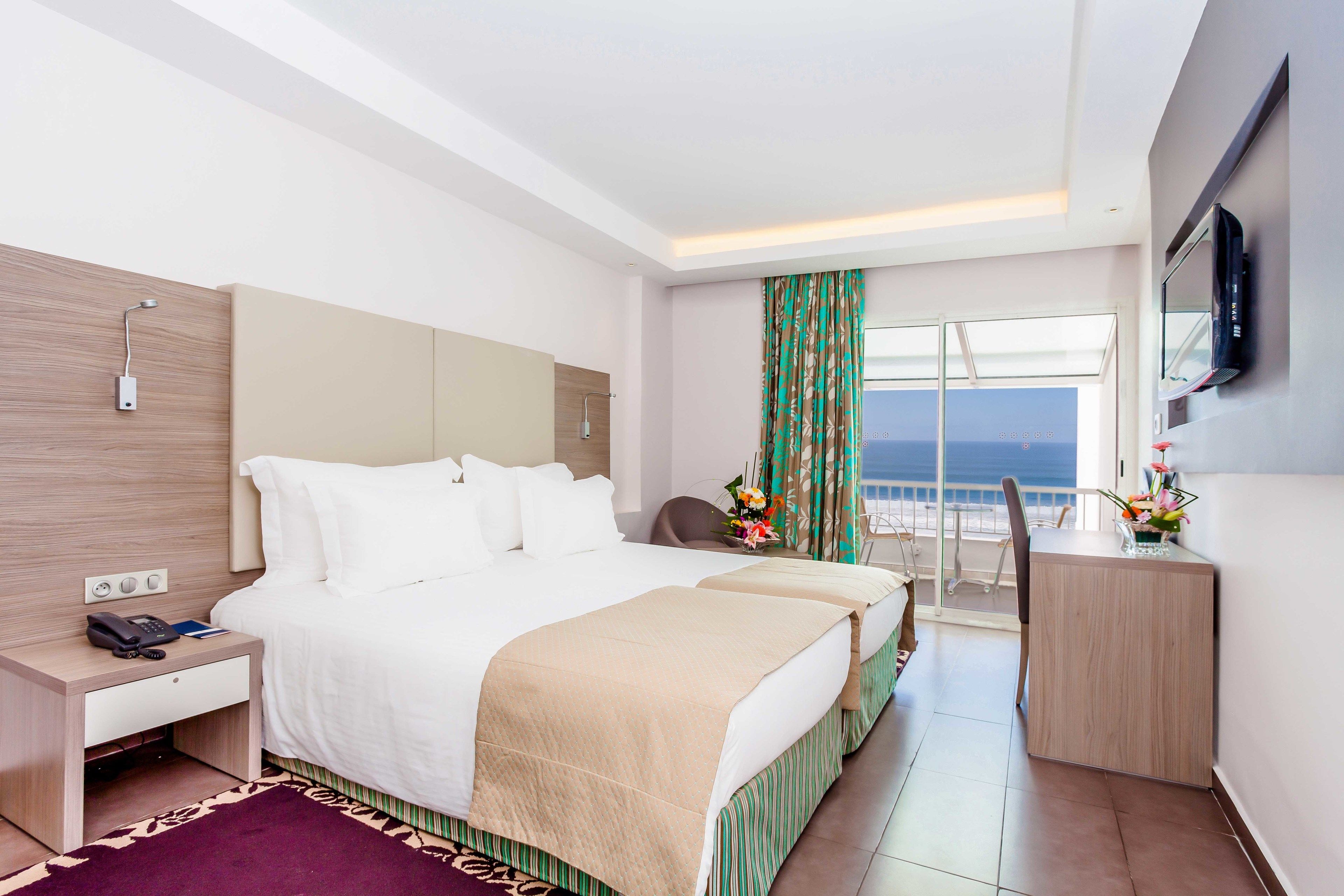 A hotel room with a large bed, a desk, a chair, a TV, and a balcony with a view.
