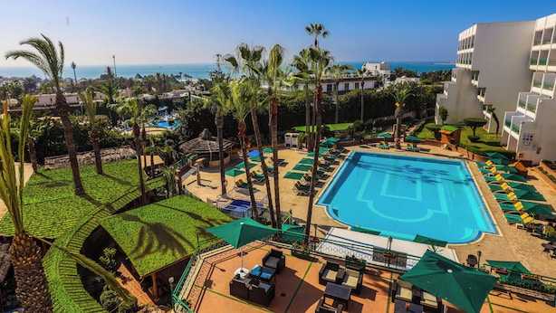 A resort with a large swimming pool, palm trees, and a view of the ocean. - Hotel Argana (Agadir)