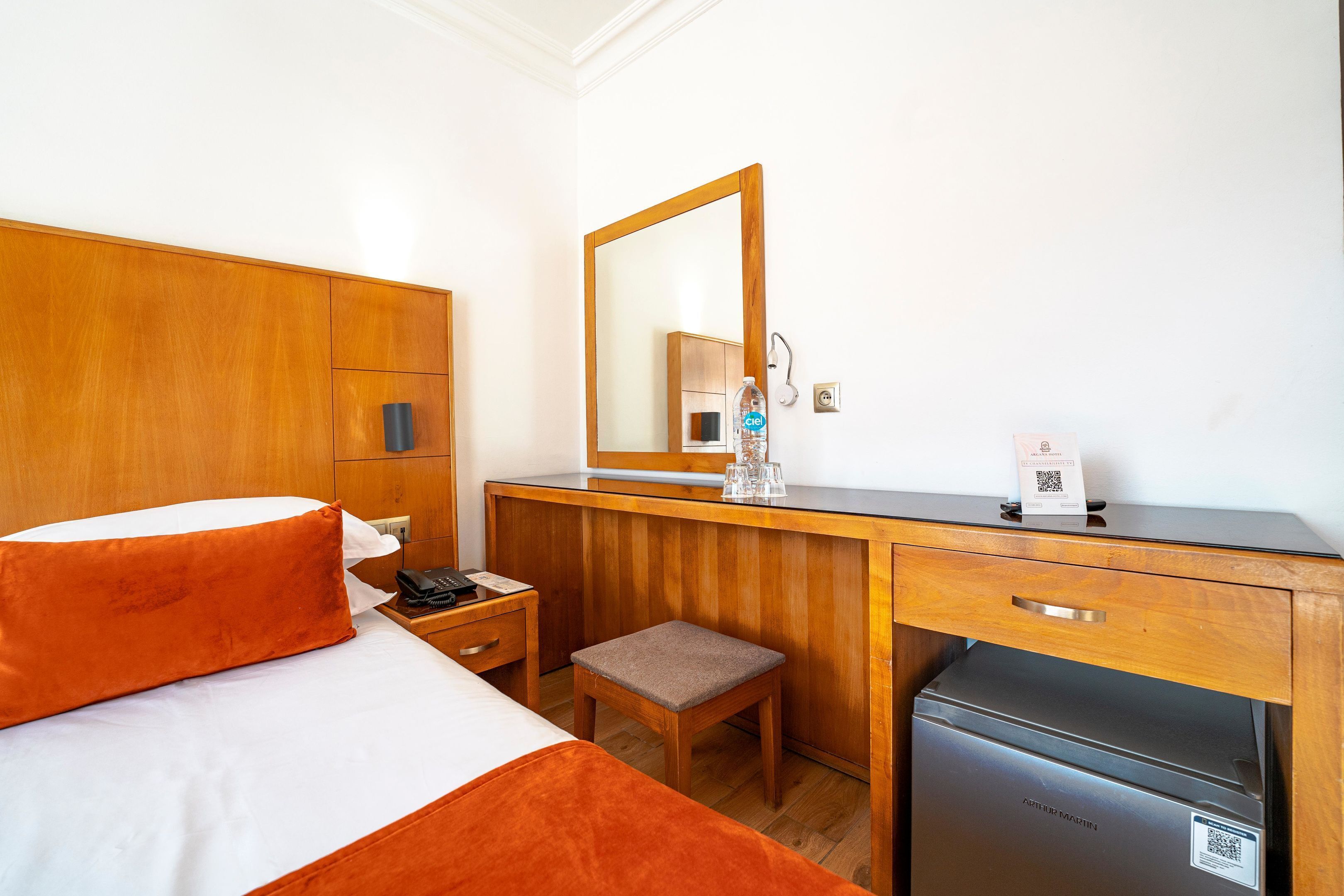 A hotel room with a bed, a wooden desk, a small stool, a refrigerator, a mirror, and a nightstand with a phone and a bottle.