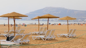 A beach with white lounge chairs and straw umbrellas, a sandy beach, and a mountainous backdrop. - Hotel Argana (Agadir)