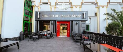 The entrance to The Art of Barber Shop with a red door, outdoor seating, and a white facade with decorative arches.