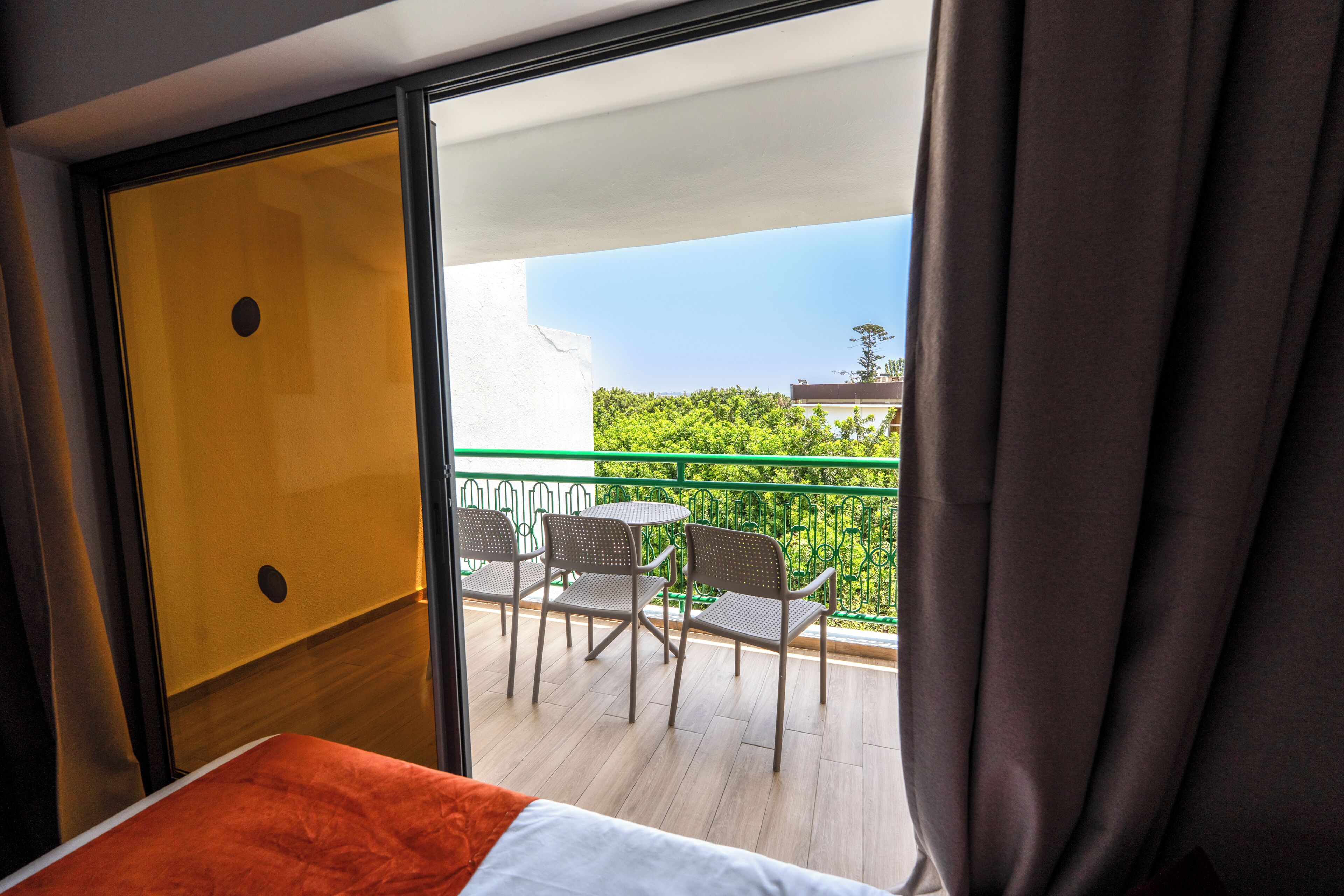 A hotel room with a balcony, a bed, and a view of greenery and buildings.