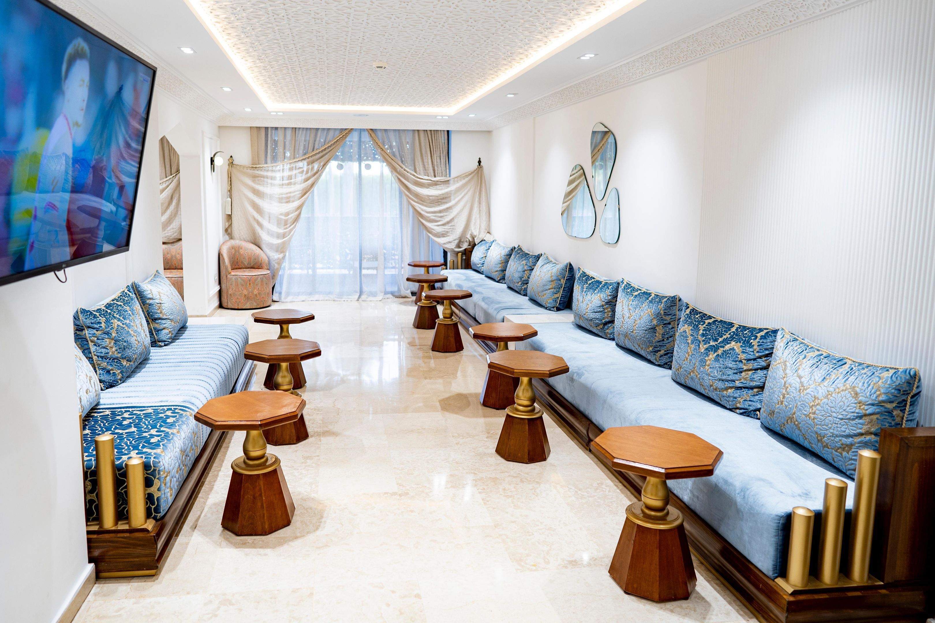 A spacious living room with a long blue sofa, multiple wooden stools, and a large flat-screen TV.