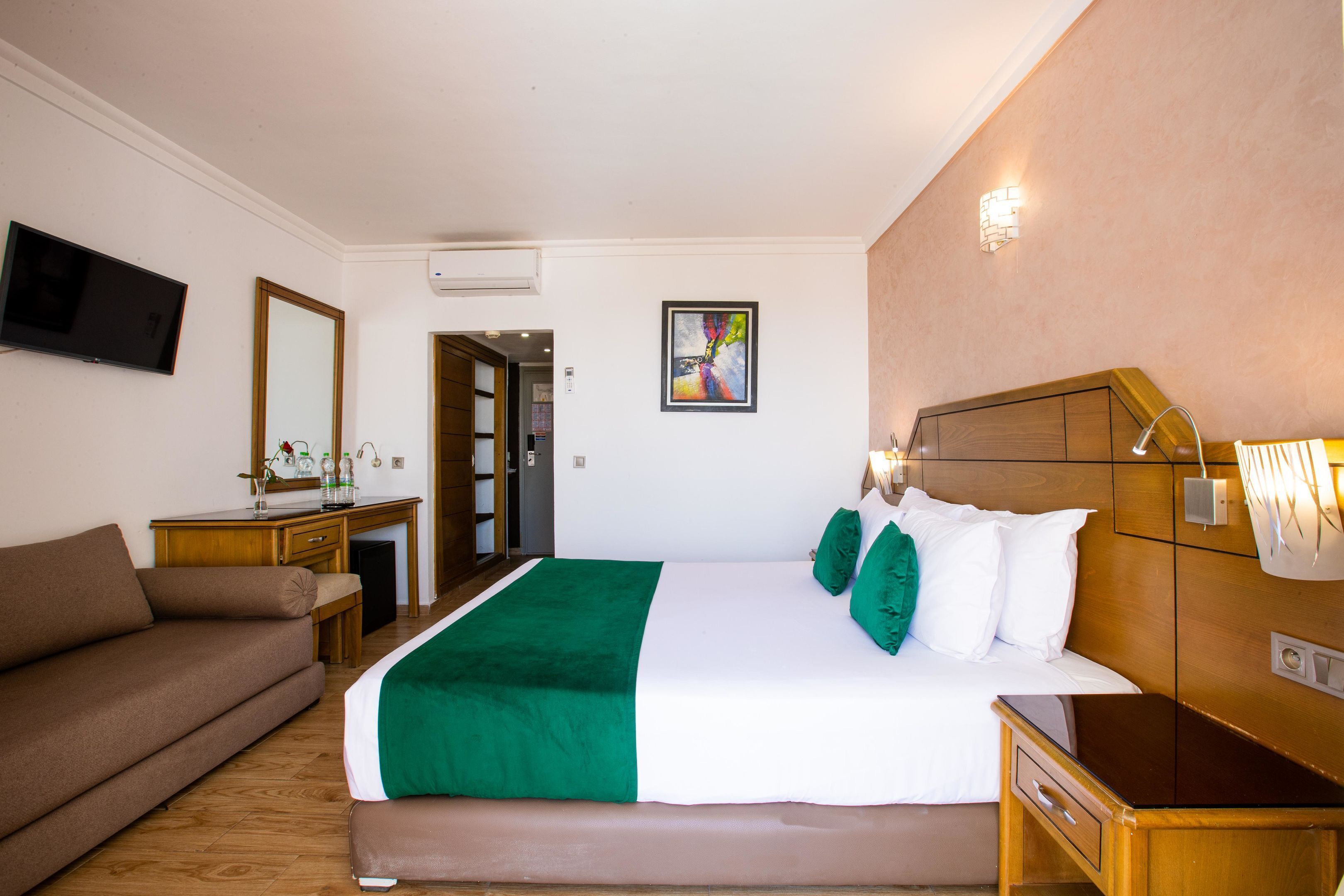 A hotel room with a bed, a sofa, a desk, a TV, and a painting on the wall.