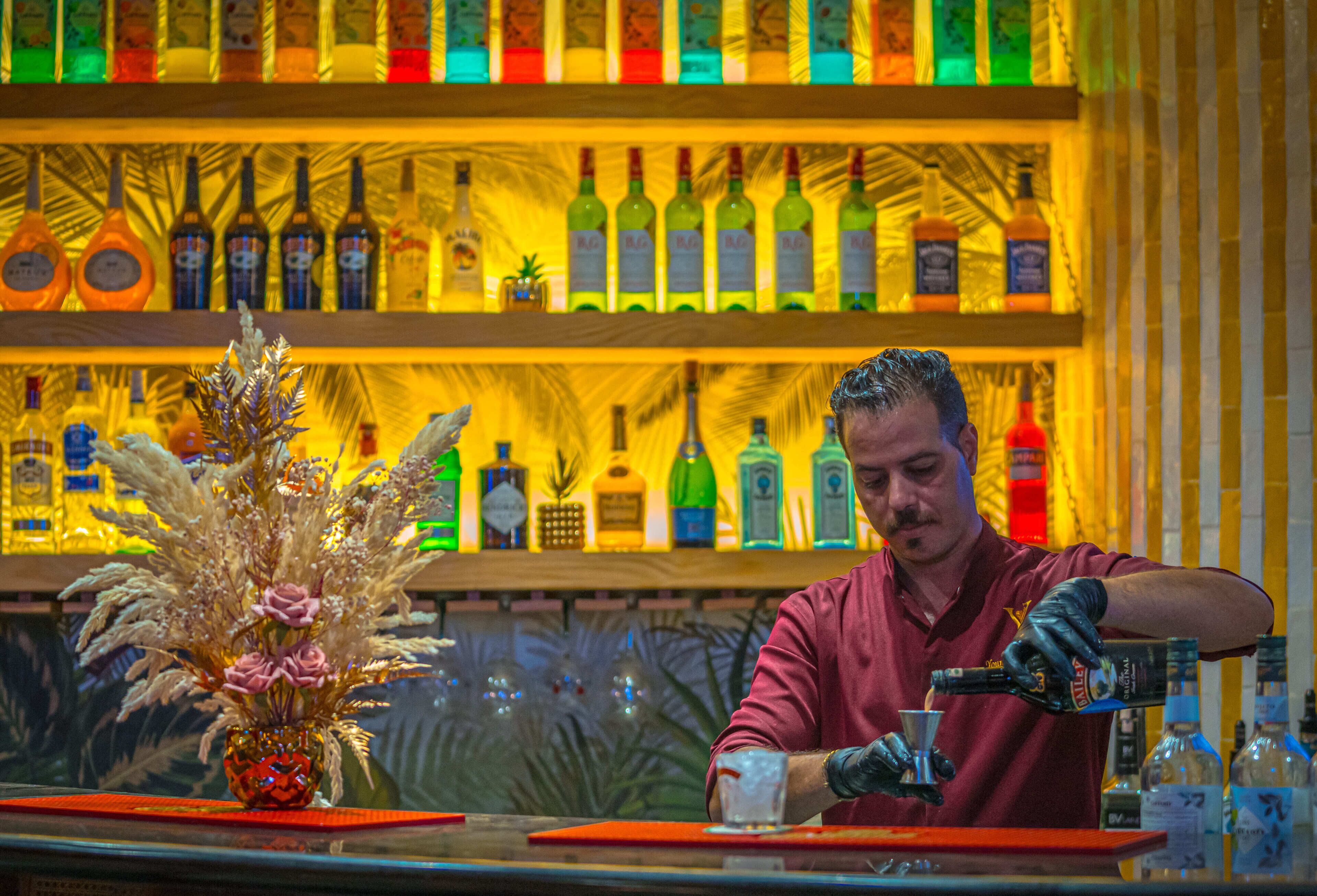 A bartender preparing a drink at a bar with shelves of liquor bottles in the background.