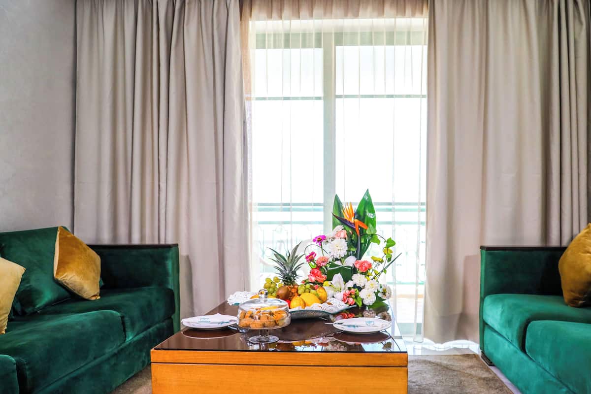 A room with two green sofas, a wooden coffee table with a fruit and flower arrangement, and a window with sheer curtains.