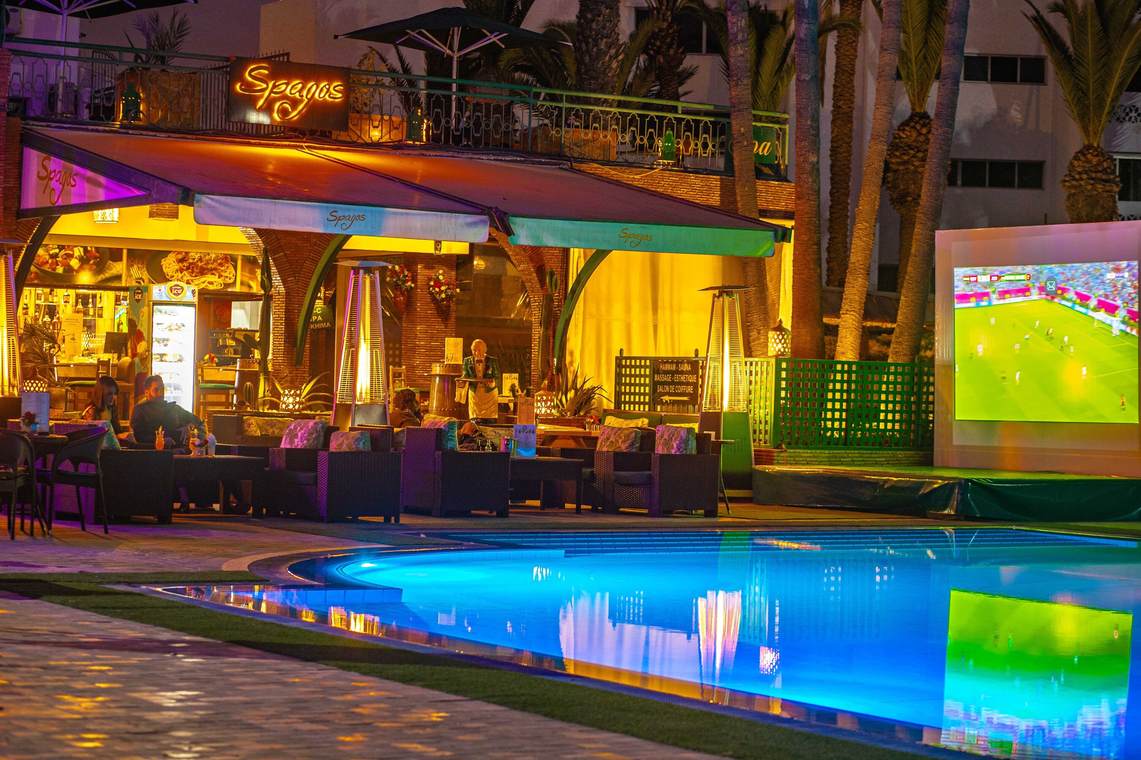 A Spagos restaurant with outdoor seating, a pool, and a large screen displaying a soccer game.