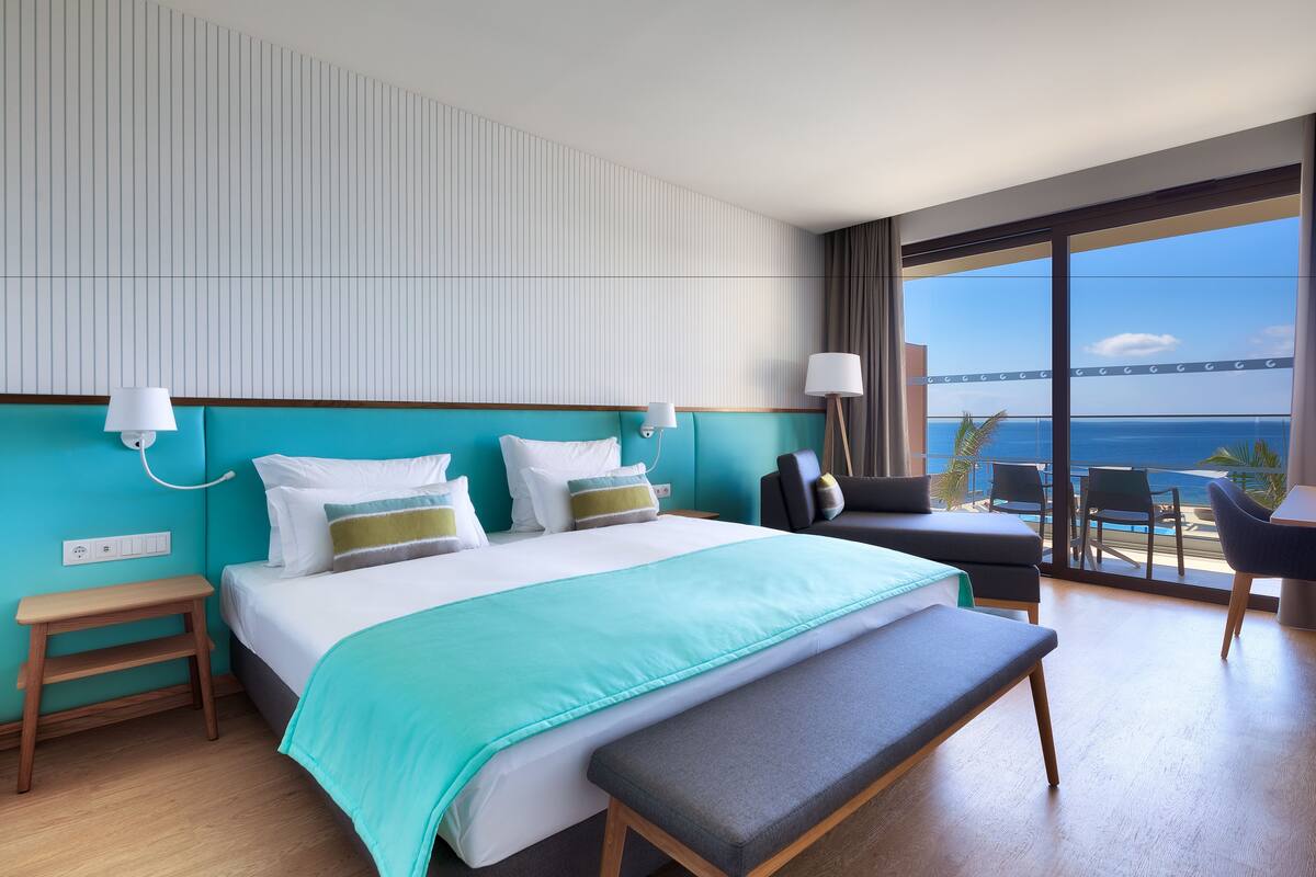 A modern hotel room with a large bed, a bench, a desk, and a view of the sea.