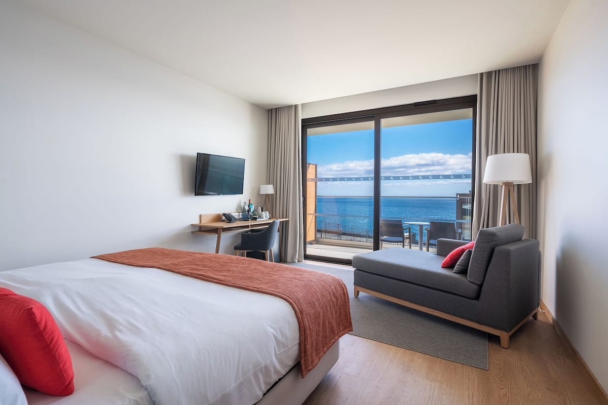 A hotel room with a bed, a desk, a sofa, and a large window with a view of the sea.