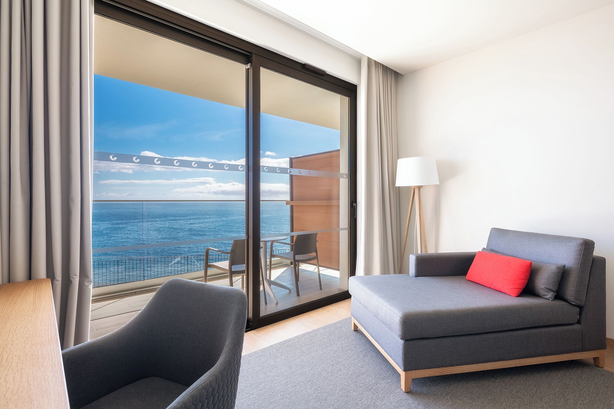 A modern hotel room with a grey sofa, a red pillow, and a view of the ocean through a large sliding glass door.