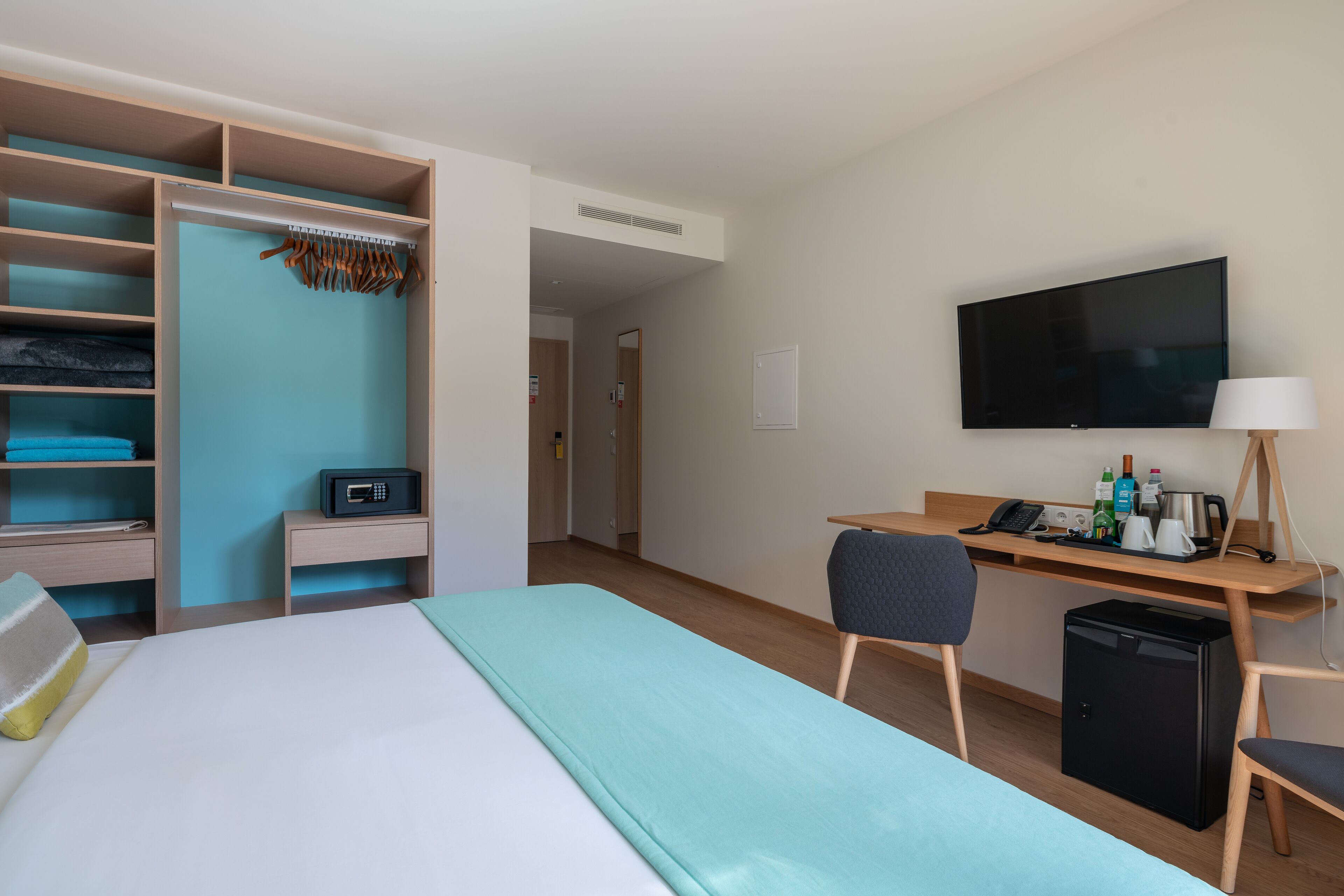A hotel room with a bed, a desk with a computer, a TV, a mini-fridge, and a wardrobe with hanging clothes.