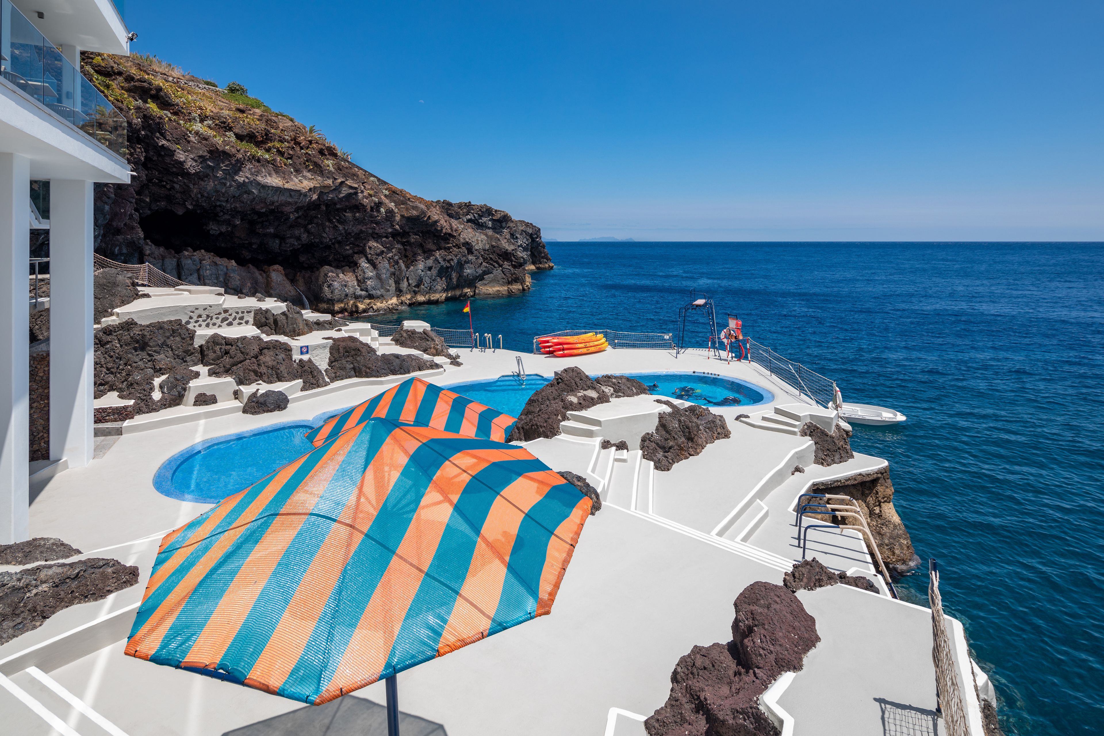A pool area with a clear view of the ocean, a rocky coastline, and a white building.