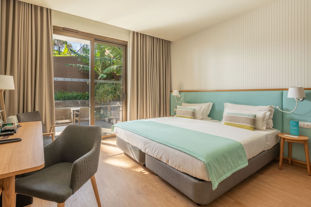 A hotel room with a large bed, a desk, a chair, and a view of greenery through the window.