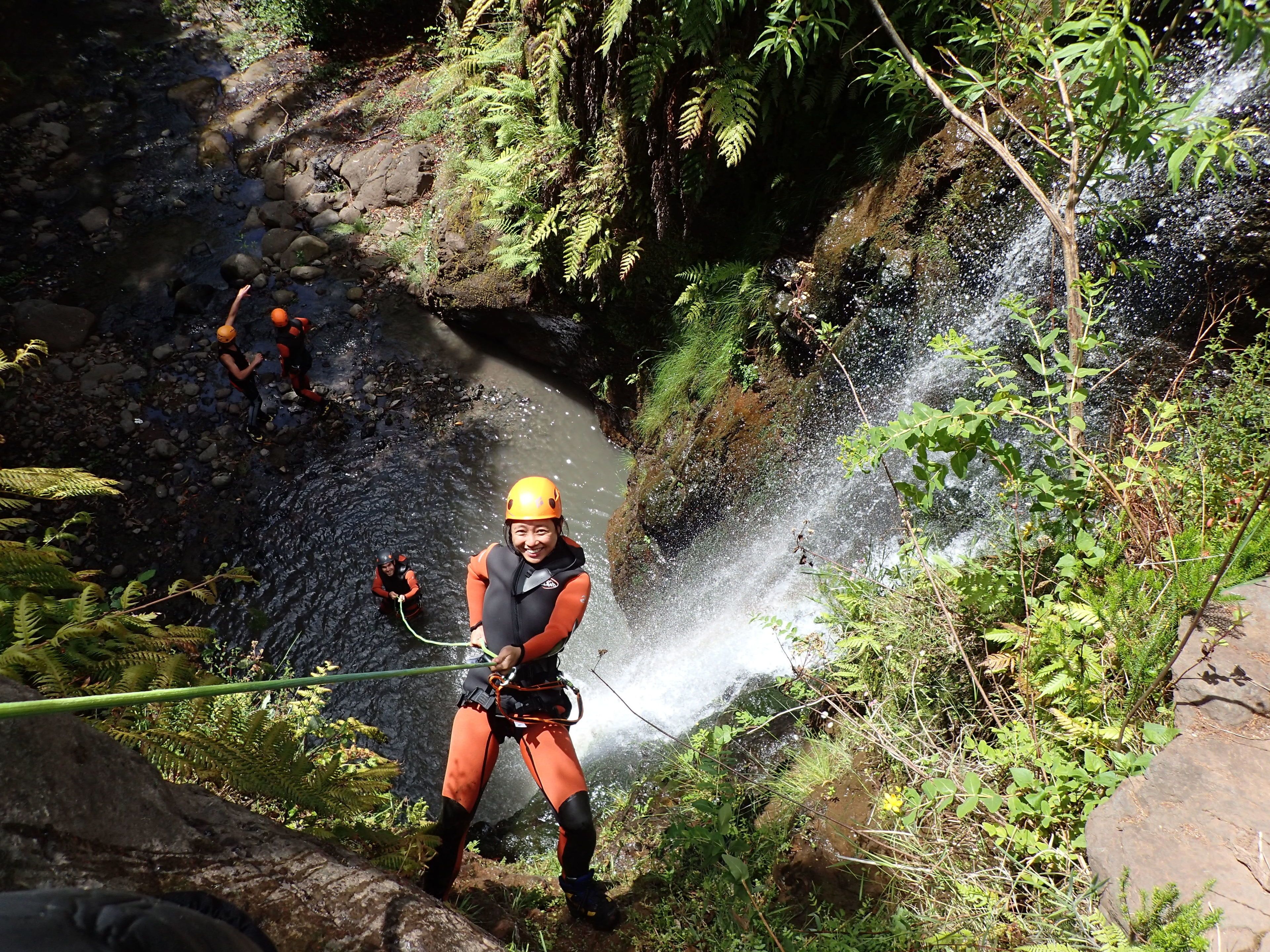 A person rappelling down a rocky cliff with a harness and helmet, surrounded by lush greenery and a waterfall.