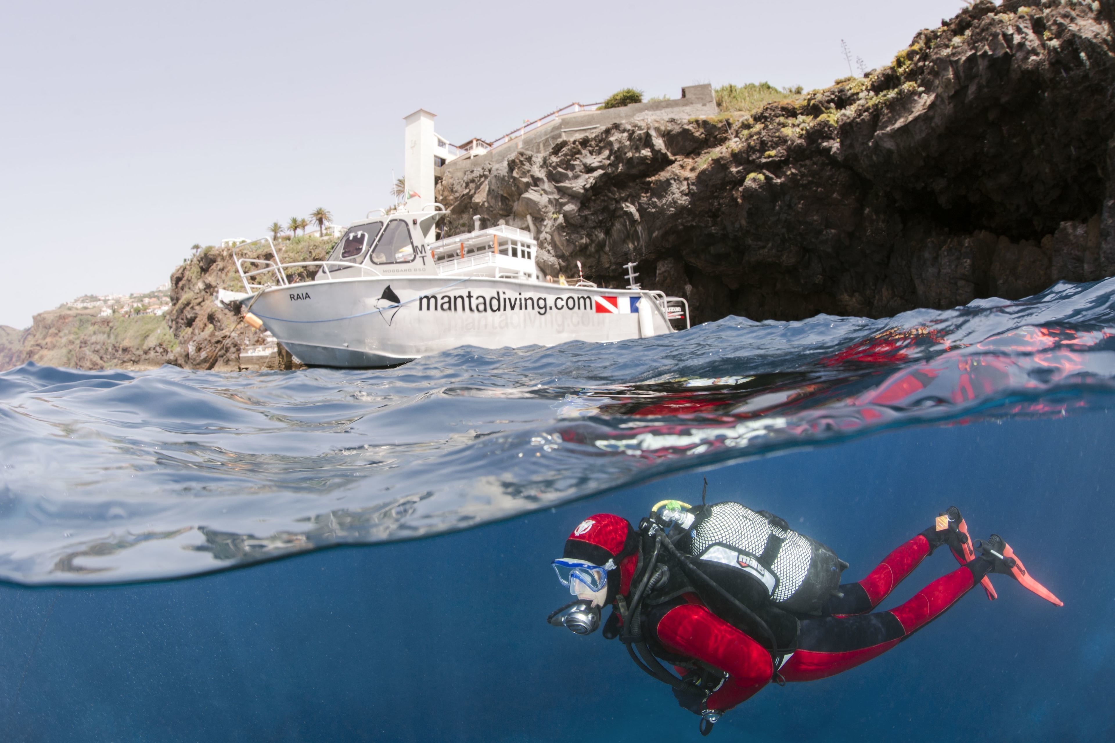 Ein Taucher in roter Ausrüstung schwimmt in der Nähe eines Bootes, auf dessen Seite die Website 'mantadiving.com' zu sehen ist, mit felsigen Klippen im Hintergrund.