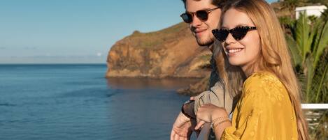 A couple standing by a railing overlooking a coastal view with a rocky cliff and greenery.