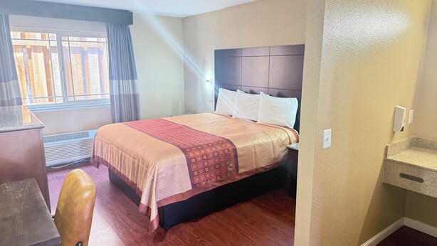 A hotel room with a bed, a desk, a chair, a window with curtains, and a wall-mounted air conditioning unit. - Valley Inn (Watsonville)