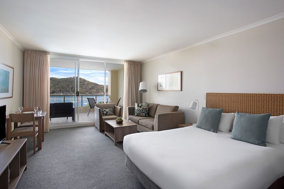 Une chambre d’hôtel avec un grand lit, un canapé, un bureau avec une chaise, une télévision et un balcon offrant une vue sur les collines.