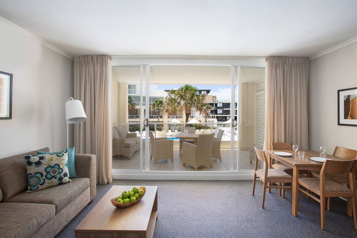 Une chambre d’hôtel avec un canapé, une table basse sur laquelle se trouve un saladier rempli de pommes, une table à manger dressée pour deux personnes, et une vue sur l’espace extérieur avec piscine.