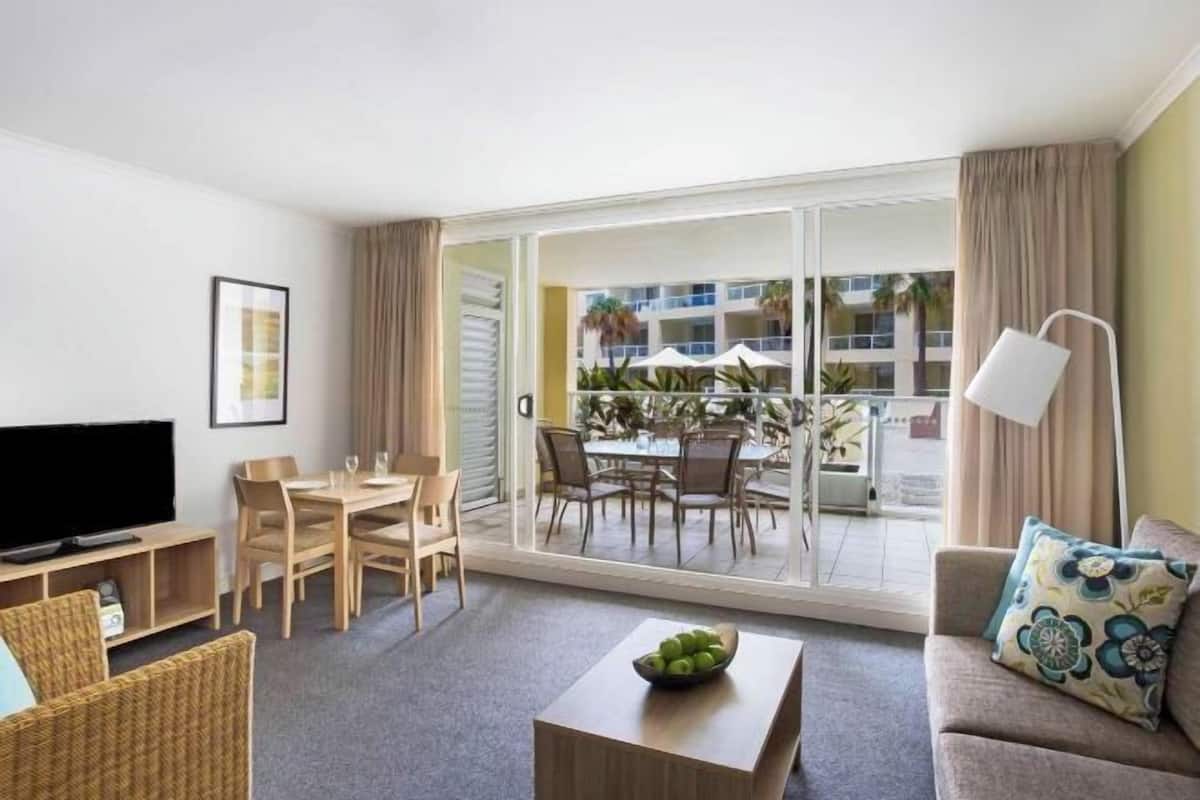 Une chambre d’hôtel dotée d’une télévision à écran plat, d’un coin repas avec une table et des chaises, d’un canapé agrémenté d’un coussin à motifs et d’un balcon avec des meubles d’extérieur et des plantes.