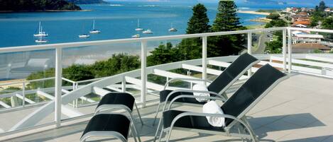 Une terrasse avec des chaises donnant sur une étendue d’eau où se trouvent des bateaux, le tout entouré de verdure et de bâtiments.