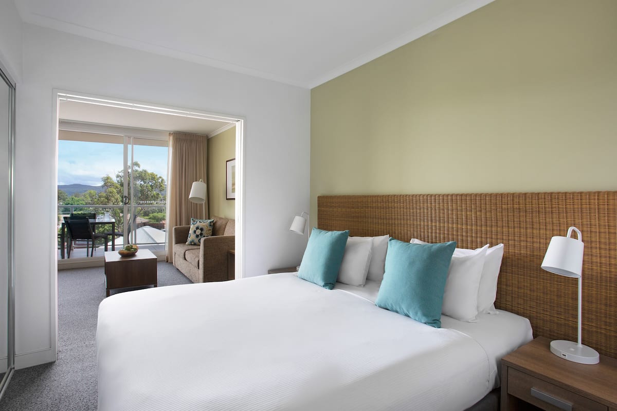Une chambre d’hôtel avec un grand lit, une table de chevet avec une lampe, une petite table avec un bol, et une vue sur l’extérieur.
