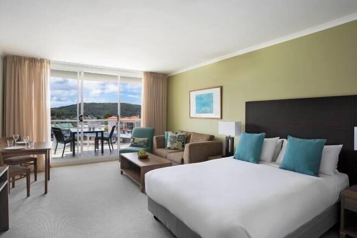 A hotel room with a large bed, a desk, a chair, a sofa, and a balcony with a table and chairs.
