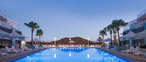 A swimming pool with lounge chairs and umbrellas, surrounded by palm trees and a mountain in the background.