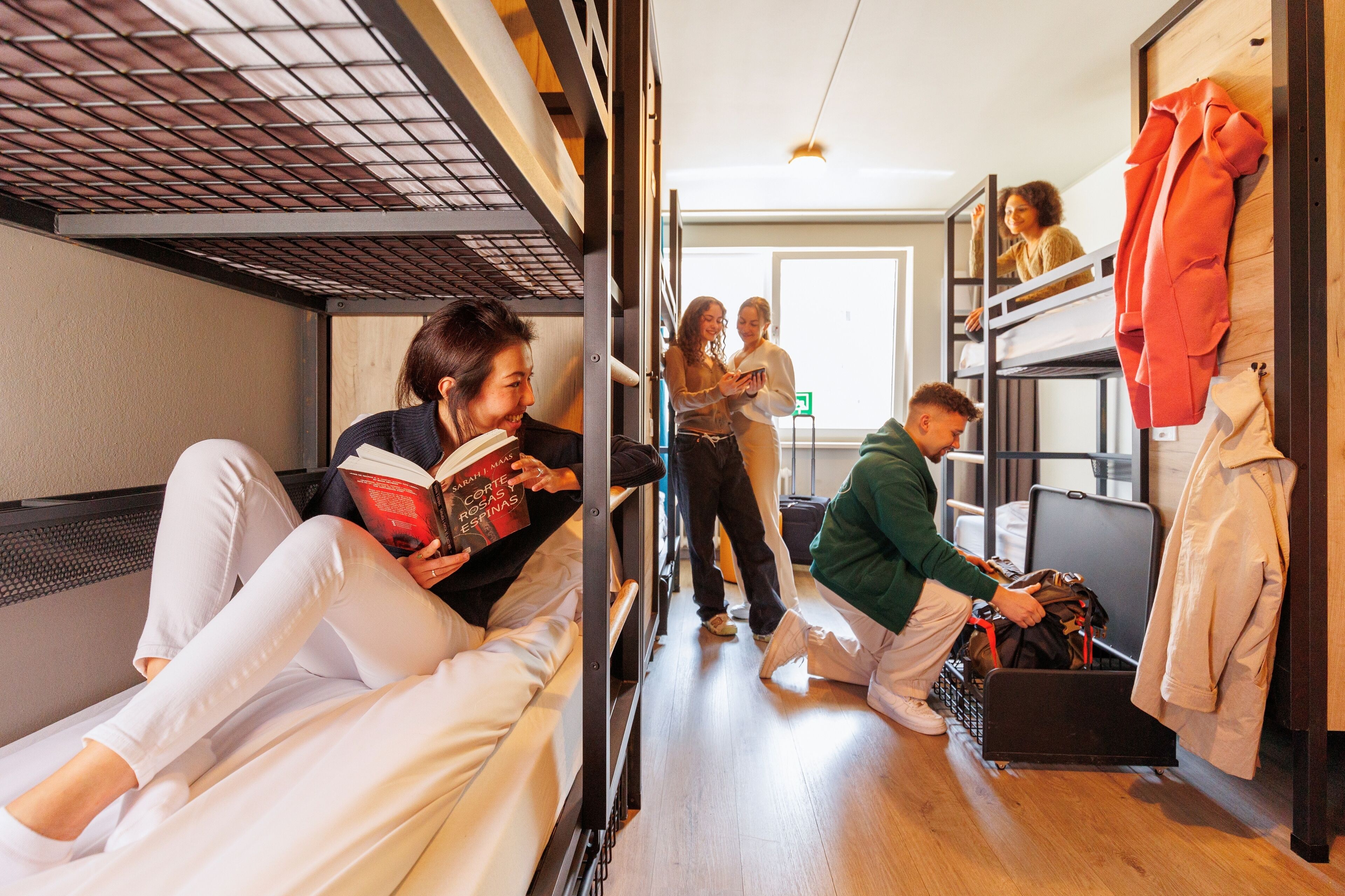Una habitación tipo dormitorio con literas, una maleta en el suelo y una persona leyendo un libro.