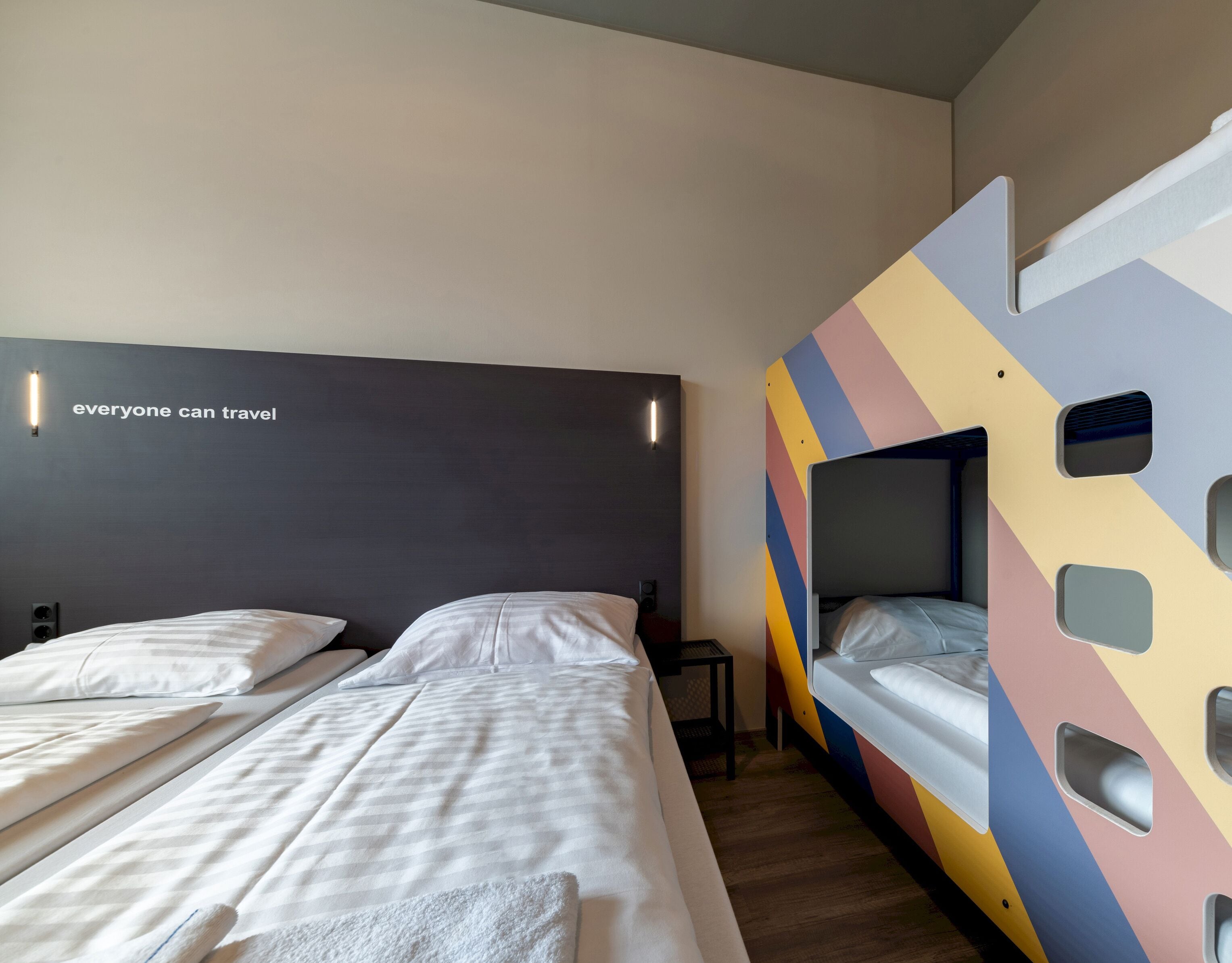 A modern hotel room with two beds, a dark headboard, a colorful geometric wall, and a motivational quote on the wall.