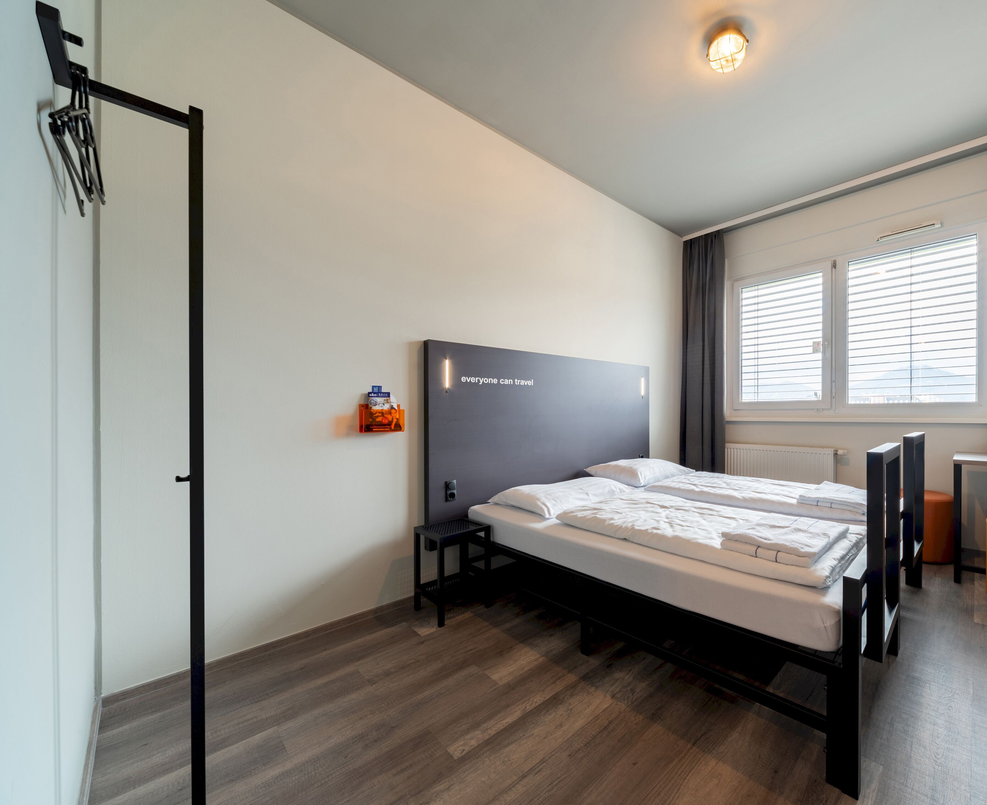 Una habitación de hotel moderna con una cama, una mesita de noche pequeña, un estante con libros en la pared, un radiador y una ventana con persianas.