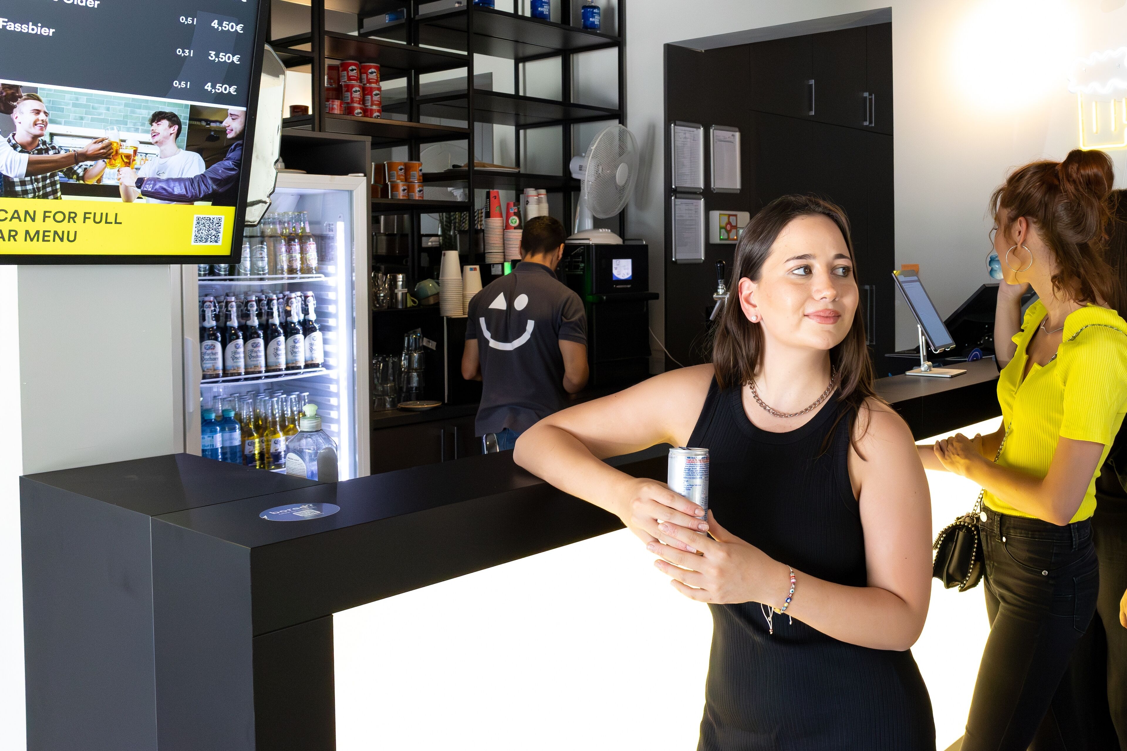 A bar area with a digital menu display, a refrigerator stocked with beverages, and a counter with a QR code for full menu access.