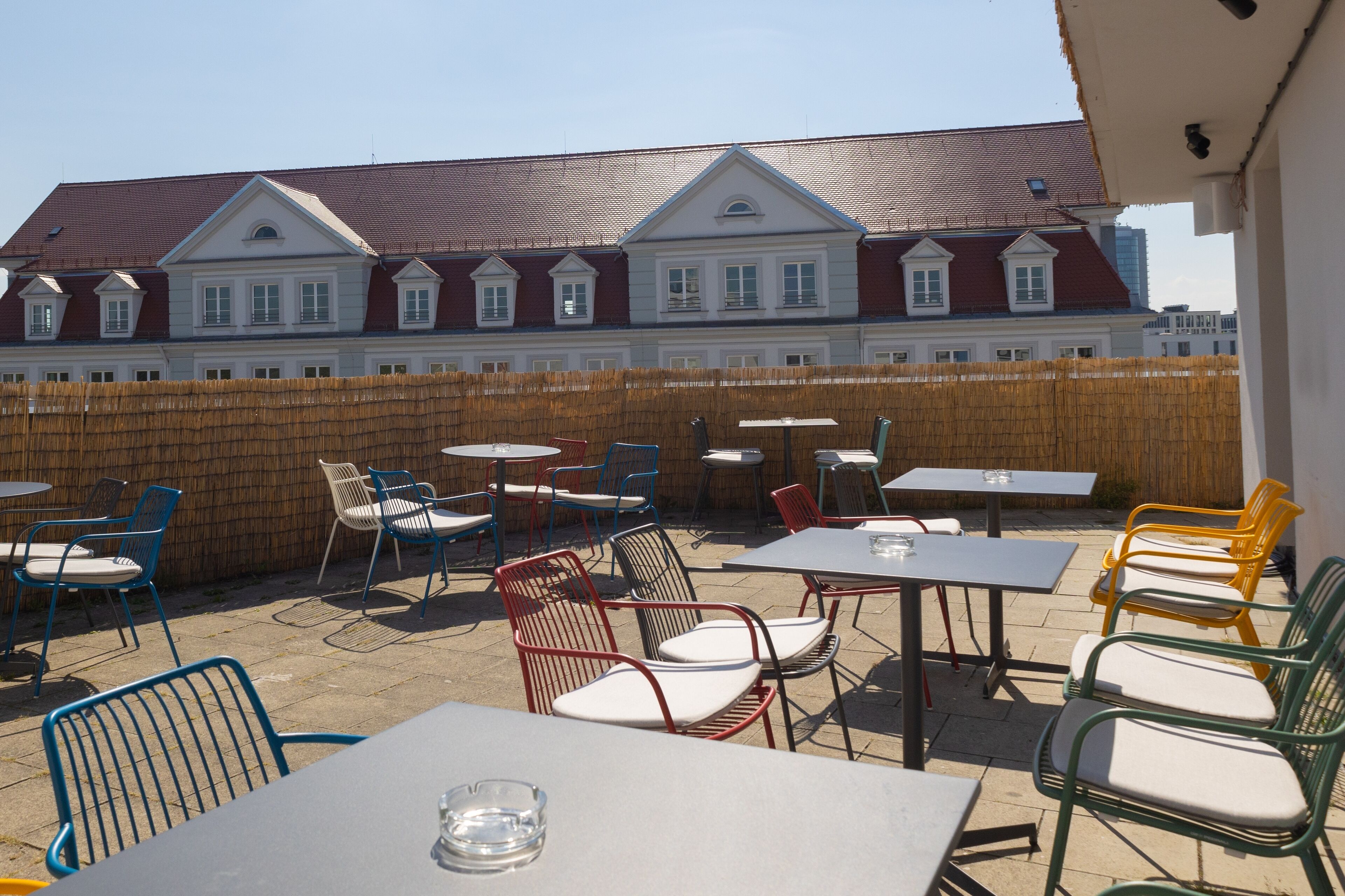 An outdoor patio area with tables and chairs, a wooden fence, and a building with a red roof in the background.