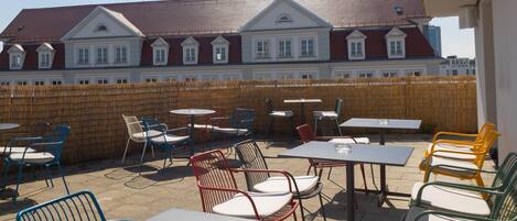 An outdoor patio area with tables and chairs, a wooden fence, and a building with a red roof in the background.