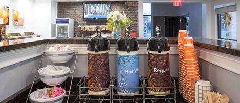 Un coin café équipé de distributeurs étiquetés « Eau chaude », « Café normal » et « Café », ainsi qu’un récipient contenant des ustensiles.