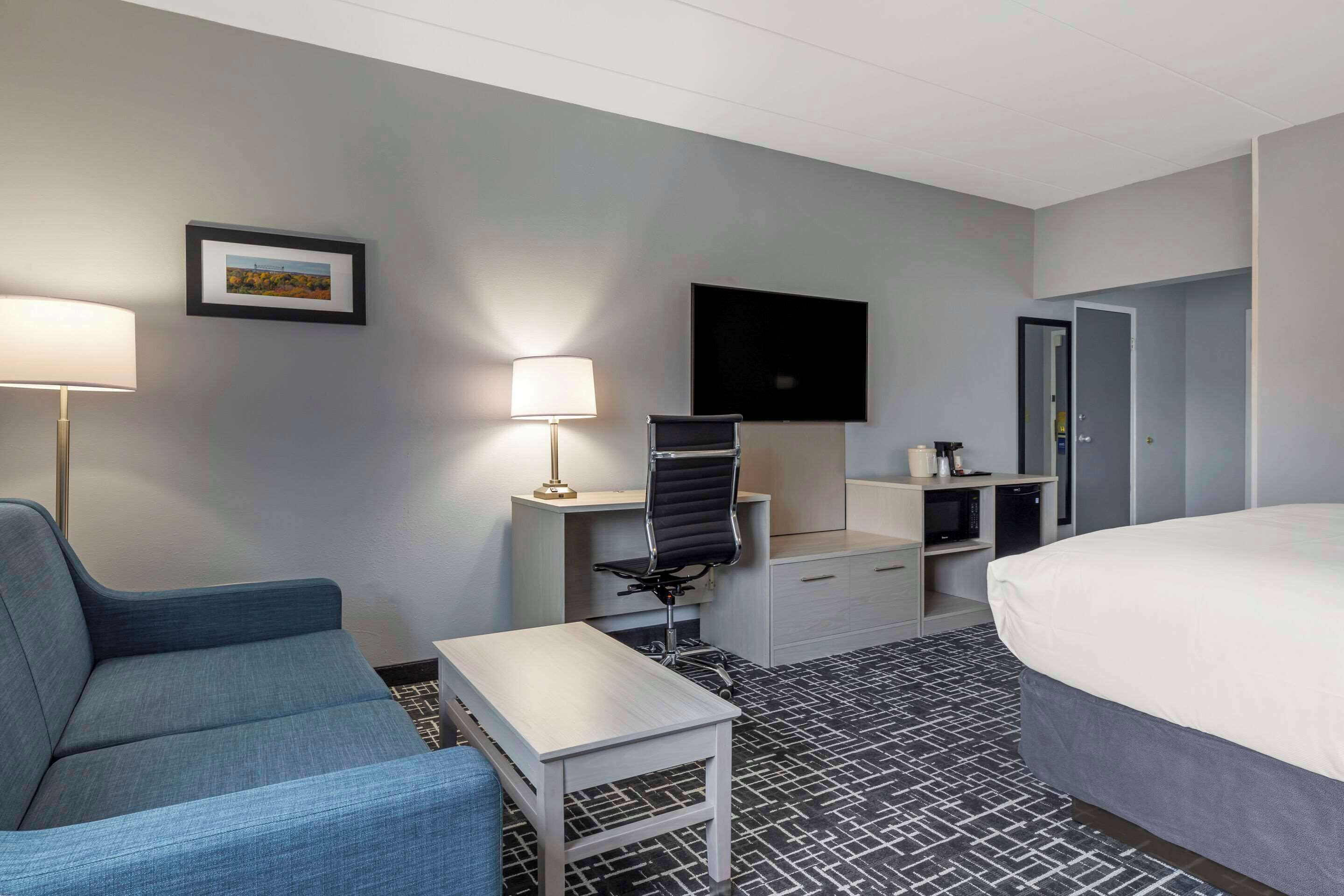 A hotel room with a bed, a desk with a chair, a TV, a lamp, and a framed picture on the wall.