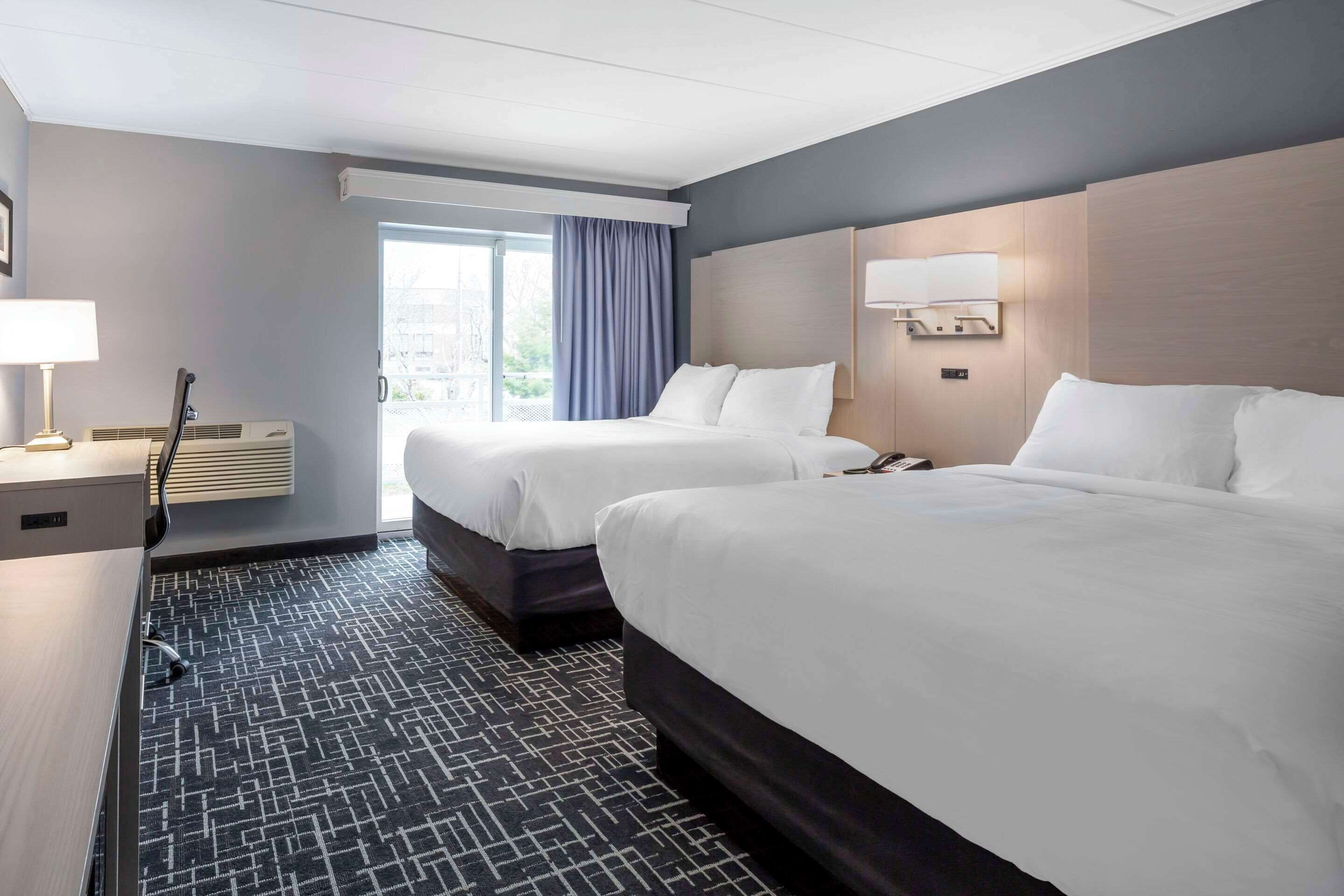 Ein Hotelzimmer mit zwei Betten, einem Schreibtisch, einer Lampe und einem Fenster mit Vorhängen.