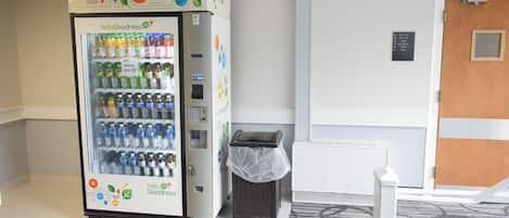A vending machine with a variety of beverages and snacks, a trash bin, and a door with a sign indicating room numbers.